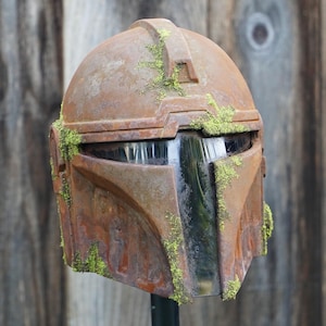 May include: A weathered, brown Mandalorian helmet with a visor, covered in patches of green moss. The helmet has a battle-worn appearance, suggesting it has seen action. The helmet is a prop or collectible.
