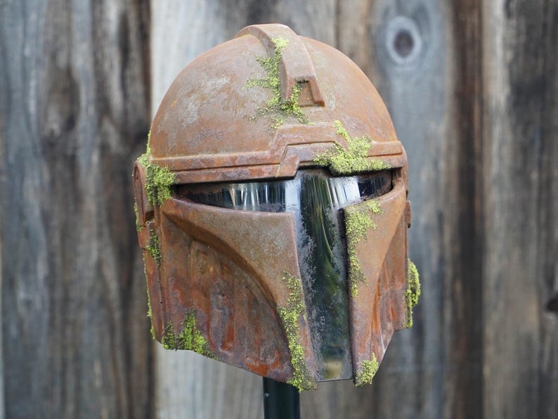 May include: A weathered, brown Mandalorian helmet with a visor, covered in patches of green moss. The helmet has a battle-worn appearance, suggesting it has seen action. The helmet is a prop or collectible.