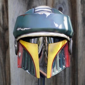K&ouml;nnte beinhalten: Ein detaillierter, verwitterter Helm mit gr&uuml;n-blauem Oberteil, gelb-roten Akzenten und einem silbernen Visier. Der Helm hat ein kampferprobtes Aussehen, was darauf hindeutet, dass es sich um eine Requisite oder ein Sammlerst&uuml;ck aus einem Science-Fiction-Franchise handelt.