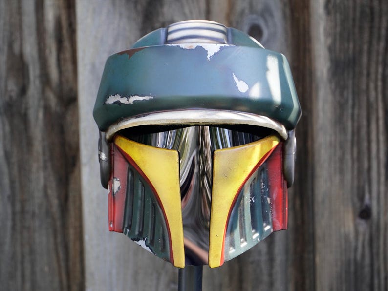 K&ouml;nnte beinhalten: Ein detaillierter, verwitterter Helm mit gr&uuml;n-blauem Oberteil, gelb-roten Akzenten und einem silbernen Visier. Der Helm hat ein kampferprobtes Aussehen, was darauf hindeutet, dass es sich um eine Requisite oder ein Sammlerst&uuml;ck aus einem Science-Fiction-Franchise handelt.