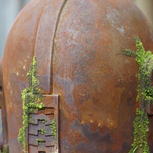 May include: Close-up of a weathered, rusty metal object, possibly a helmet or sculpture. The surface is a mottled brown with patches of green moss growing on the side. The object has a rounded top and geometric details.