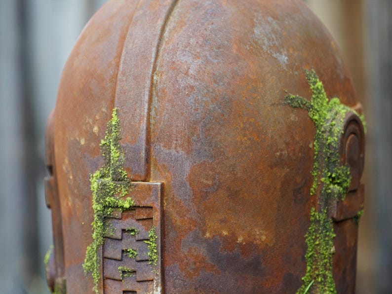 May include: Close-up of a weathered, rusty metal object, possibly a helmet or sculpture. The surface is a mottled brown with patches of green moss growing on the side. The object has a rounded top and geometric details.