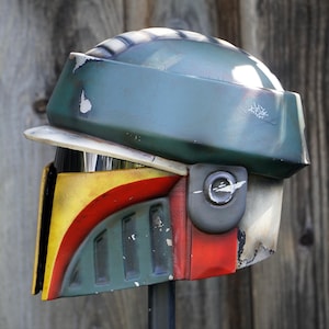 K&ouml;nnte beinhalten: Ein verwitterter Helm mit einem gr&uuml;n, gelb, rot und wei&szlig; farbigen Farbschema. Der Helm hat ein Visier und ein seitlich angebrachtes Ger&auml;t. Das Design erinnert an eine bekannte Science-Fiction-Figur.