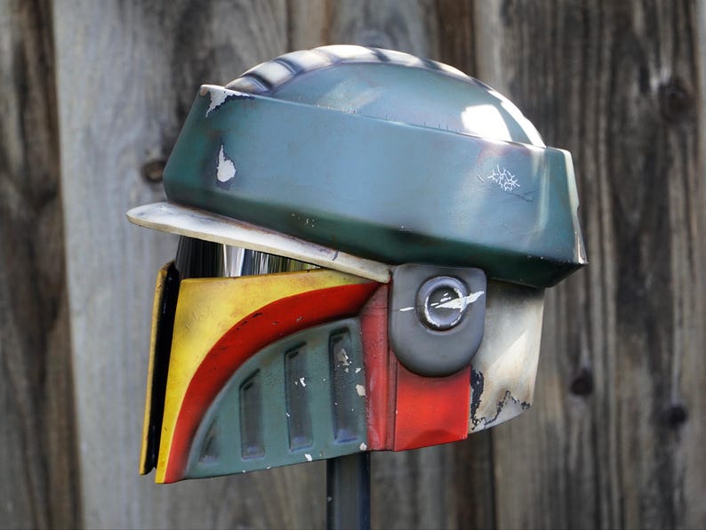 K&ouml;nnte beinhalten: Ein verwitterter Helm mit einem gr&uuml;n, gelb, rot und wei&szlig; farbigen Farbschema. Der Helm hat ein Visier und ein seitlich angebrachtes Ger&auml;t. Das Design erinnert an eine bekannte Science-Fiction-Figur.