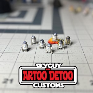 Peut inclure: Plusieurs figurines miniatures de droïdes R2-D2 argentés et dorés sont disposées sur une surface quadrillée. Le logo "SKYGUY MINI ARTOO DETOO CUSTOMS" en rouge et blanc est également visible.