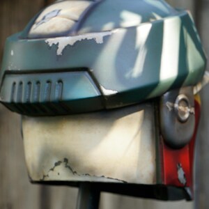 K&ouml;nnte beinhalten: Ein verwitterter, gr&uuml;n-wei&szlig;er Helm mit Visier und L&uuml;ftungsschlitzen. Der Helm hat ein abgenutztes Aussehen mit sichtbaren Lackabplatzungen, was auf eine Vintage- oder kampferprobte &Auml;sthetik hindeutet. Der Helm ist ein Requisit oder Kost&uuml;mteil.