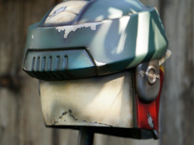 K&ouml;nnte beinhalten: Ein verwitterter, gr&uuml;n-wei&szlig;er Helm mit Visier und L&uuml;ftungsschlitzen. Der Helm hat ein abgenutztes Aussehen mit sichtbaren Lackabplatzungen, was auf eine Vintage- oder kampferprobte &Auml;sthetik hindeutet. Der Helm ist ein Requisit oder Kost&uuml;mteil.