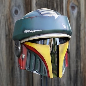 K&ouml;nnte beinhalten: Ein verwitterter, gr&uuml;n-blauer Helm mit gelben und roten Akzenten. Der Helm hat ein Visier und ist wahrscheinlich ein Kost&uuml;mteil oder Sammlerst&uuml;ck, m&ouml;glicherweise aus einem Science-Fiction-Franchise.
