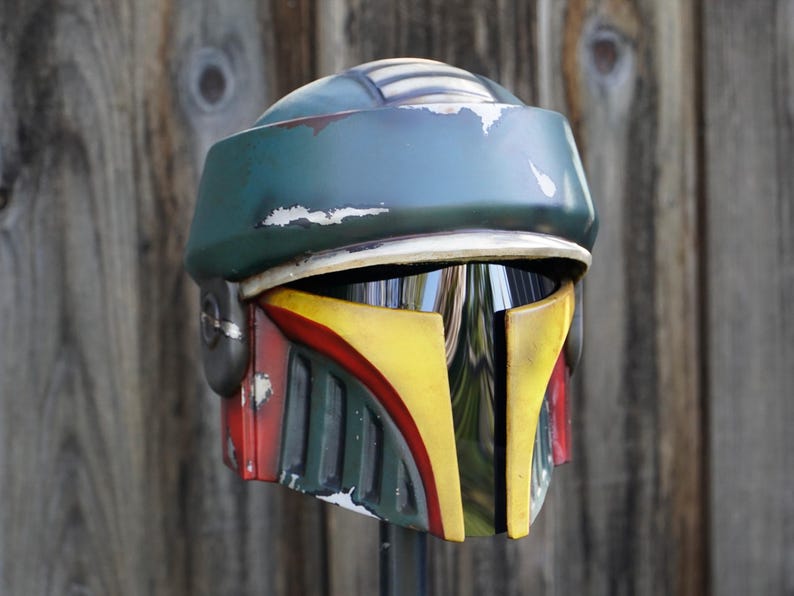 K&ouml;nnte beinhalten: Ein verwitterter, gr&uuml;n-blauer Helm mit gelben und roten Akzenten. Der Helm hat ein Visier und ist wahrscheinlich ein Kost&uuml;mteil oder Sammlerst&uuml;ck, m&ouml;glicherweise aus einem Science-Fiction-Franchise.