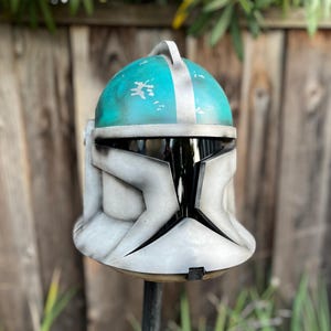 May include: A white and teal clone trooper helmet with a black visor. The helmet has a weathered look with white paint splatters on the teal surface.