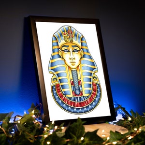 May include: A framed print of a colorful illustration of an Egyptian pharaoh's mask. The mask is blue, gold, and red with a detailed design. The pharaoh's face is depicted with a serious expression.