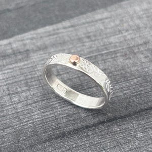 May include: A silver ring with an engraved floral pattern and a small, round, rose gold accent. The ring is set against a gray background, showcasing its intricate design and metallic shine.
