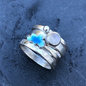 May include: A set of four silver rings stacked on top of each other. The top ring has a clear stone, the second ring has a blue enamel cloud, the third ring has a swirl design, and the bottom ring has a floral design.