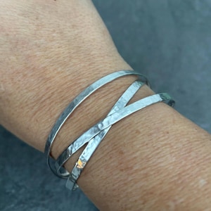 May include: A silver-toned bracelet with a hammered texture. The bracelet features three bands, two of which cross over each other. The jewelry is worn on a wrist and has a simple, yet elegant design.