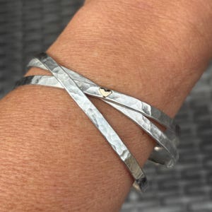May include: A silver-toned, hammered-metal bracelet with three overlapping bands. A small gold-colored heart charm is visible on one band. The bracelet has a textured surface and a simple, elegant design, suitable for everyday wear.