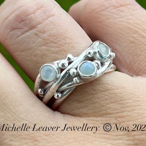 May include: A silver ring with three moonstone gems set in a woven band. The ring is worn on a person's finger.