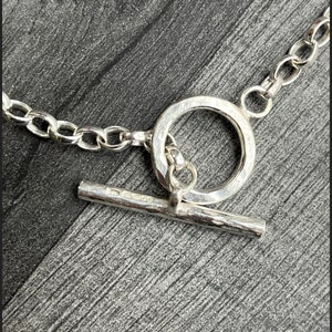 May include: Silver chain necklace with a toggle clasp. The chain is made of small, linked circles. The clasp is a round ring with a bar that slides through it.