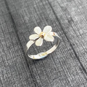 May include: A silver ring featuring a flower-shaped design with white petals and a small gold-colored center. The band is made of polished silver and has a smooth, rounded finish. The flower sits prominently on top of the band, creating a simple, clean look.