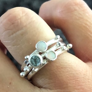 Peut inclure: Une bague en argent avec trois bandes superposées, chacune avec de petites perles et une pierre précieuse. Les pierres précieuses sont une aquamarine bleu clair et deux opales blanches.