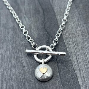 May include: Silver necklace featuring a toggle clasp and a pendant. The pendant is a textured, circular shape with a small gold heart at its centre. The chain is made of interconnected oval links.