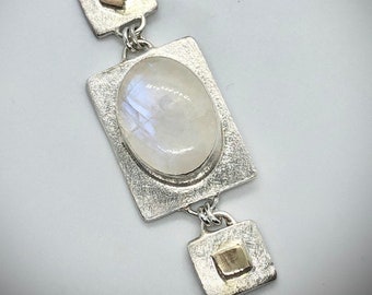 May include: A silver necklace with a moonstone pendant and two small gold accents. The pendant is a rectangular shape with a textured surface. The moonstone is oval shaped and has a white iridescent sheen.