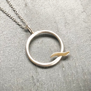 May include: A silver necklace with a circle pendant featuring a gold accent. The pendant is a simple circle with a small gold wave design.