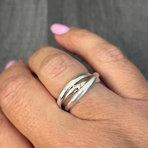 May include: A silver ring with a gold heart-shaped accent. The ring has a layered design with three bands that intertwine. The ring is worn on a finger with a pink nail polish.