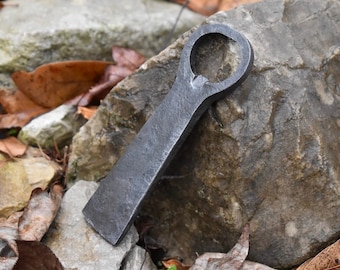 Può includere: Un apribottiglie forgiato a mano in metallo con una finitura rustica. L'apribottiglie ha la forma di una piccola ascia con un'apertura arrotondata per aprire le bottiglie.