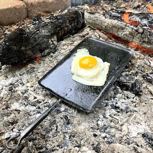 Könnte beinhalten: Ein Spiegelei auf einer schwarzen Metallplatte über einem Lagerfeuer. Das Ei hat ein hellgelbes Eigelb und weiße Eiweiß. Die Platte hat einen langen Griff. Das Feuer ist von Asche und verkohltem Holz umgeben.