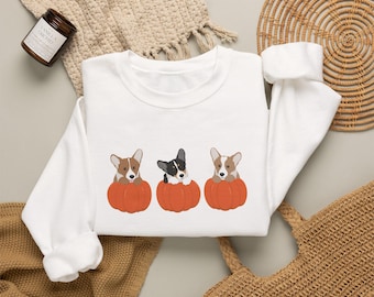 May include: A white long-sleeve sweatshirt featuring a graphic print of three Corgi dogs, each sitting inside a bright orange pumpkin. The dogs are depicted in different color variations, including tan and white, and black and white.