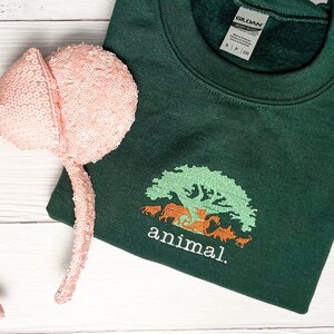 May include: A green sweatshirt with a white embroidered tree and animals, and the text "animal."  A pink sequined headband with a bow is also in the image.