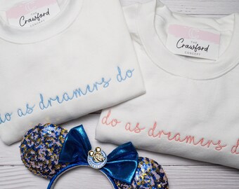 Op de afbeelding: Twee witte sweatshirts met de tekst "do as dreamers do" geborduurd in blauw en roze. De sweatshirts zijn gevouwen en liggen op een houten oppervlak. Een paar blauwe en gouden, met pailletten bezette Minnie Mouse-oren bevinden zich op de voorgrond.