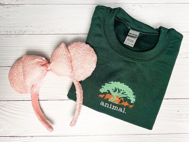May include: A green sweatshirt with a white embroidered tree and animals, and the text "animal."  A pink sequined headband with a bow is also in the image.