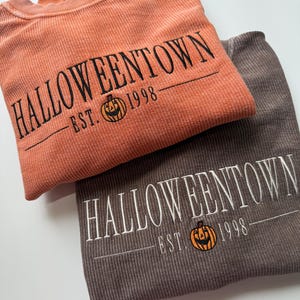 May include: Two corded sweatshirts, one orange and one brown, are folded and stacked. The orange sweatshirt has black embroidered text that reads "Halloweentown" with "Est. 1998" and a jack-o'-lantern graphic. The brown sweatshirt has white embroidery.