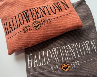 May include: Two corded sweatshirts, one orange and one brown, are folded and stacked. The orange sweatshirt has black embroidered text that reads "Halloweentown" with "Est. 1998" and a jack-o'-lantern graphic. The brown sweatshirt has white embroidery.