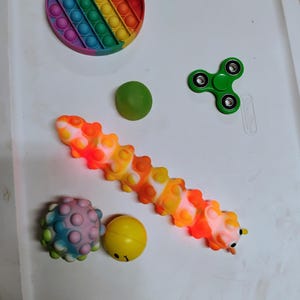 May include: A selection of fidget toys in various shapes and colours. Includes a rainbow pop-it, a green fidget spinner, a caterpillar-shaped pop-it, a spiky ball, a green ball, and a yellow ball with a face.