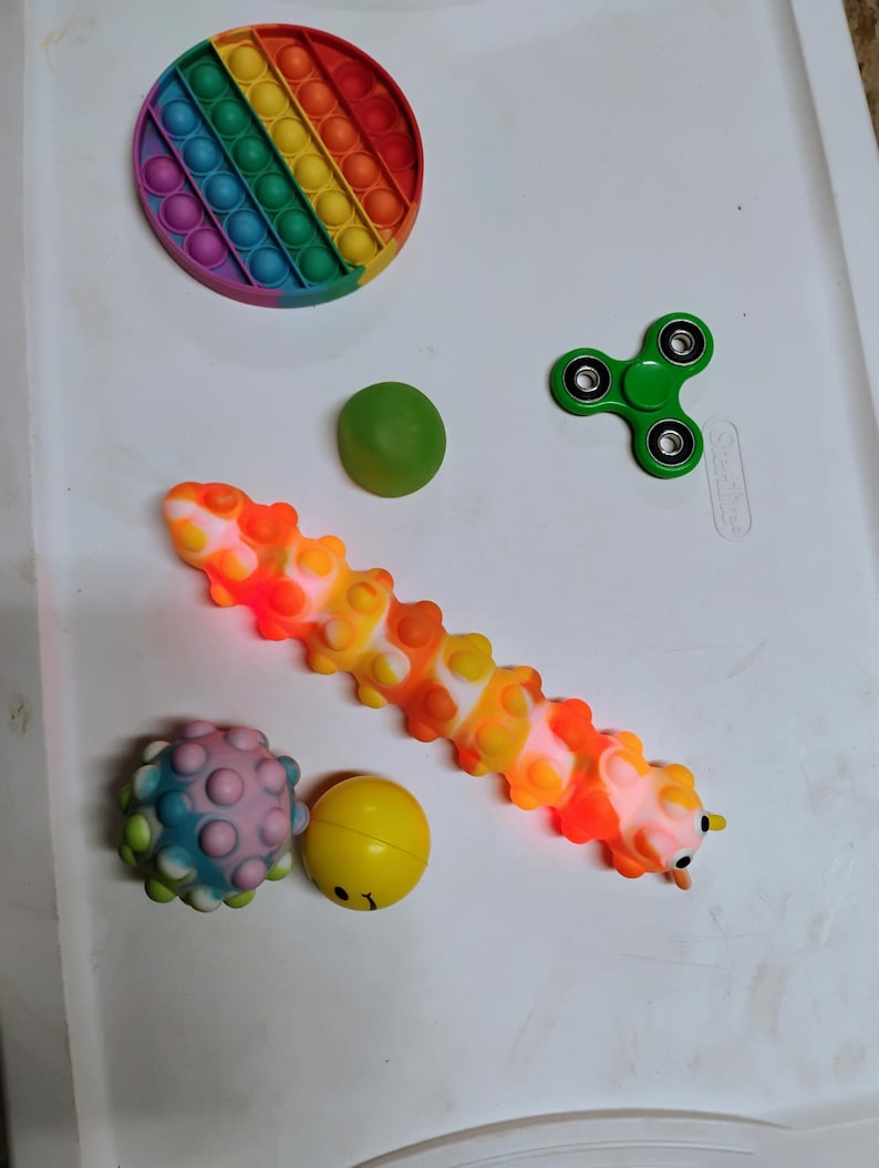 May include: A selection of fidget toys in various shapes and colours. Includes a rainbow pop-it, a green fidget spinner, a caterpillar-shaped pop-it, a spiky ball, a green ball, and a yellow ball with a face.
