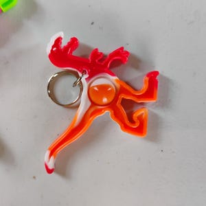 May include: A colorful lizard-shaped keychain in red, orange, and white. The keychain has a silver ring for attaching keys. The design is a stylized reptile with a playful, cartoonish appearance.