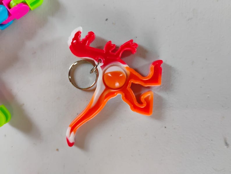 May include: A colorful lizard-shaped keychain in red, orange, and white. The keychain has a silver ring for attaching keys. The design is a stylized reptile with a playful, cartoonish appearance.