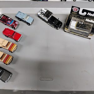 May include: A collection of die-cast model pickup trucks in various colors, including red, black, tan, and blue. One truck is packaged in a box with "Holley" branding. The trucks are arranged on a gray plastic tray.