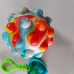 May include: A colorful, spherical sensory toy with a textured surface, featuring a mix of red, blue, yellow, and white. A green clip and a blue and green heart-shaped fidget toy are attached.