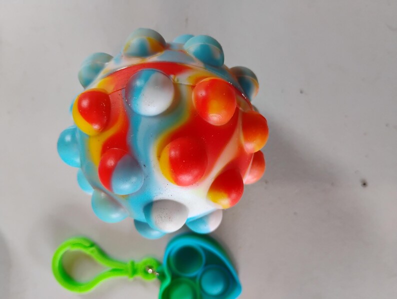 May include: A colorful, spherical sensory toy with a textured surface, featuring a mix of red, blue, yellow, and white. A green clip and a blue and green heart-shaped fidget toy are attached.