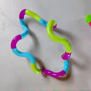 May include: A colorful fidget toy in a wavy shape, featuring segments of blue, green, and magenta. Another smaller toy with pink, blue, and green segments is also visible. The toys are designed for tactile stimulation and stress relief.