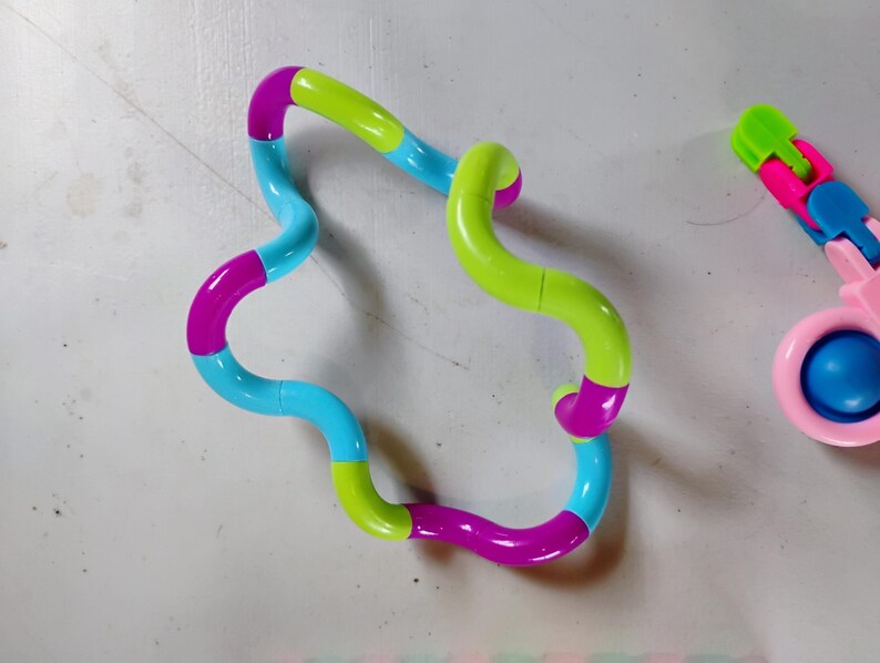 May include: A colorful fidget toy in a wavy shape, featuring segments of blue, green, and magenta. Another smaller toy with pink, blue, and green segments is also visible. The toys are designed for tactile stimulation and stress relief.