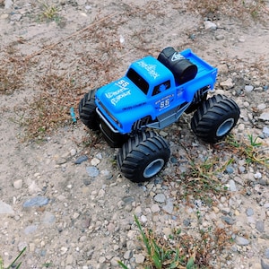 May include: A blue toy monster truck with oversized black tires and silver rims. The truck features the number 55, the text Cross-Country, and a black spare tire mounted in the truck bed. The body has white graphic designs on the hood and roof.