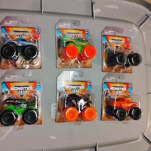 May include: Six Monster Jam toy trucks on display. Each truck is packaged on a cardboard backing with the Monster Jam logo. The trucks are various colors, including blue, green, and orange, with large black wheels.