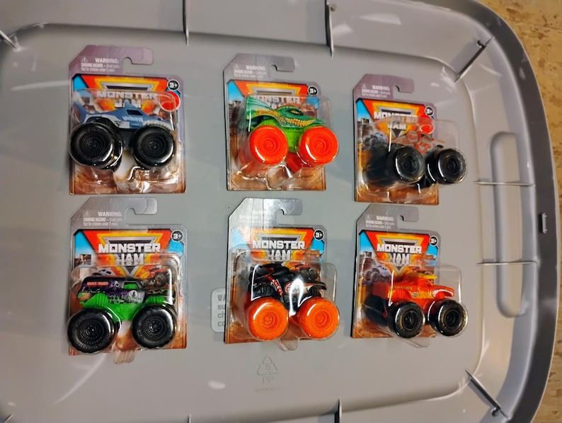 May include: Six Monster Jam toy trucks on display. Each truck is packaged on a cardboard backing with the Monster Jam logo. The trucks are various colors, including blue, green, and orange, with large black wheels.