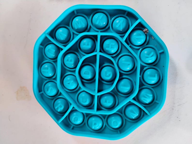May include: A turquoise octagonal fidget toy with alphabet letters. The toy has a series of circular bubbles, each with a letter of the alphabet. The letters are in a white font. The toy is made of a soft, flexible material.