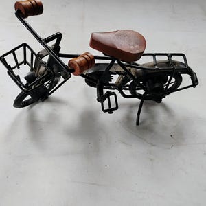 Può includere: Un modello di bicicletta in miniatura costruito in filo metallico nero, dotato di sellino in legno marrone, manopole del manubrio in legno marrone, un cestino anteriore in filo metallico, un portapacchi posteriore e ruote nere.