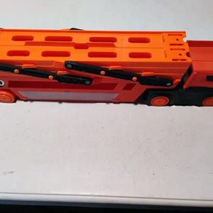 May include: A bright orange and black toy car hauler truck featuring a two-level trailer design with multiple rectangular cutouts. The side of the trailer displays the number 50 in a white circle, and the truck has black wheels and a black cab base.